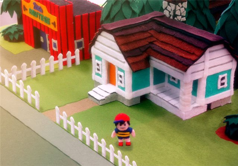 Toy figure stands by green house with white fence; another red building labeled Drug Store in background. Bright, handcrafted paper style.
